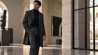 Tall man in charcoal tailoring in a contemporary Milan gallery with travertine arches.