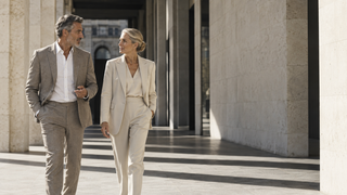 Two senior colleagues in tailored neutral suits walking through contemporary Milan architecture.