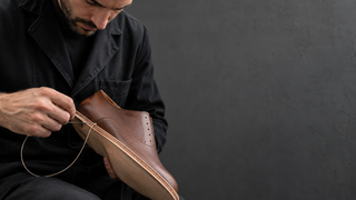 Artisan on the left hand-stitching the welt and sole of an unbranded leather dress shoe in a modern atelier.