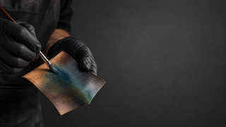 Gloved artisan on the left applying a blue-green patina to an unbranded piece of leather in a modern studio.