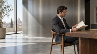 Man in a charcoal suit reviewing a plain sketchbook in a contemporary Italian atelier with brushed-steel architecture.