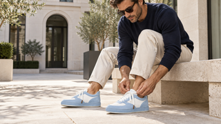 Man tying pale blue and white suede-and-leather sneakers while seated in a modern Italian courtyard.