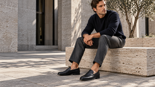 Man wearing black pebbled leather penny loafers while seated on a travertine bench in a modern Italian courtyard.